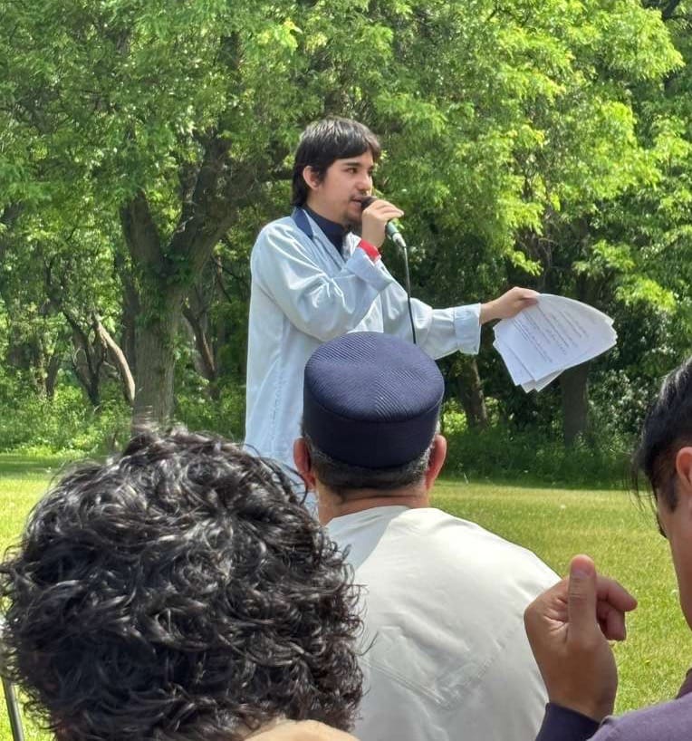 A young man in a light blue robe speaks into a microphone outdoors, holding papers, while attendees listen in the foreground.