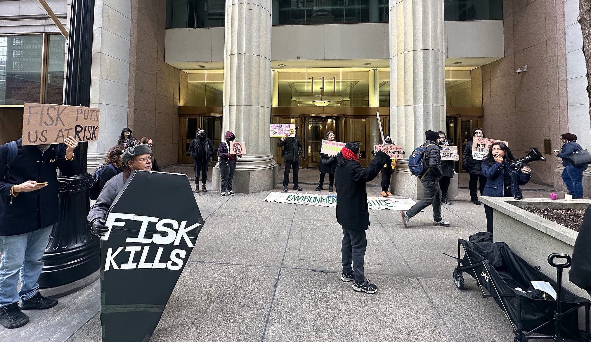 A wide shot of protesters spread across the entrance of a government building. A coffin-shaped sign reading "Fisk Kills" is visible in the foreground. Demonstrators hold signs and an "Environmental Justice" banner is laid on the ground.