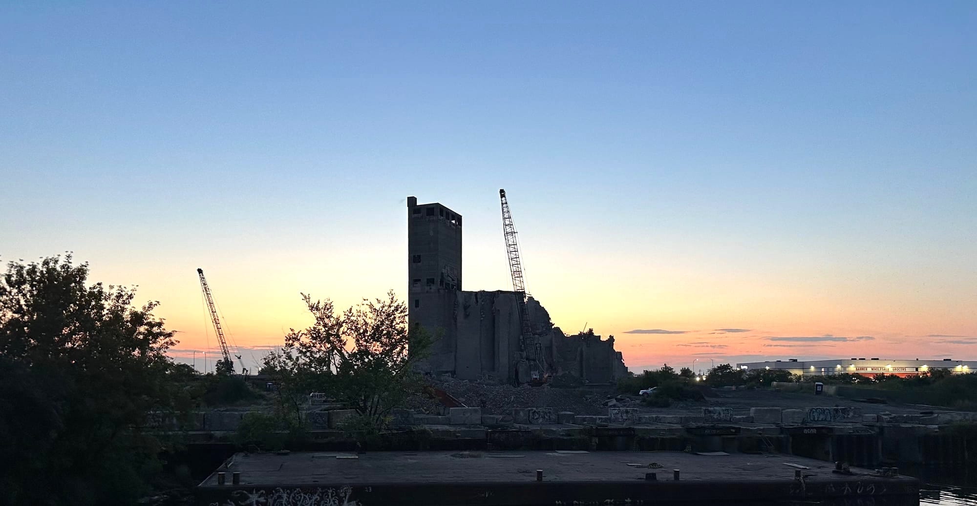 A silhouette of the partially demolished Fisk coal plant tower at sunset, flanked by two large cranes against an orange and blue sky.