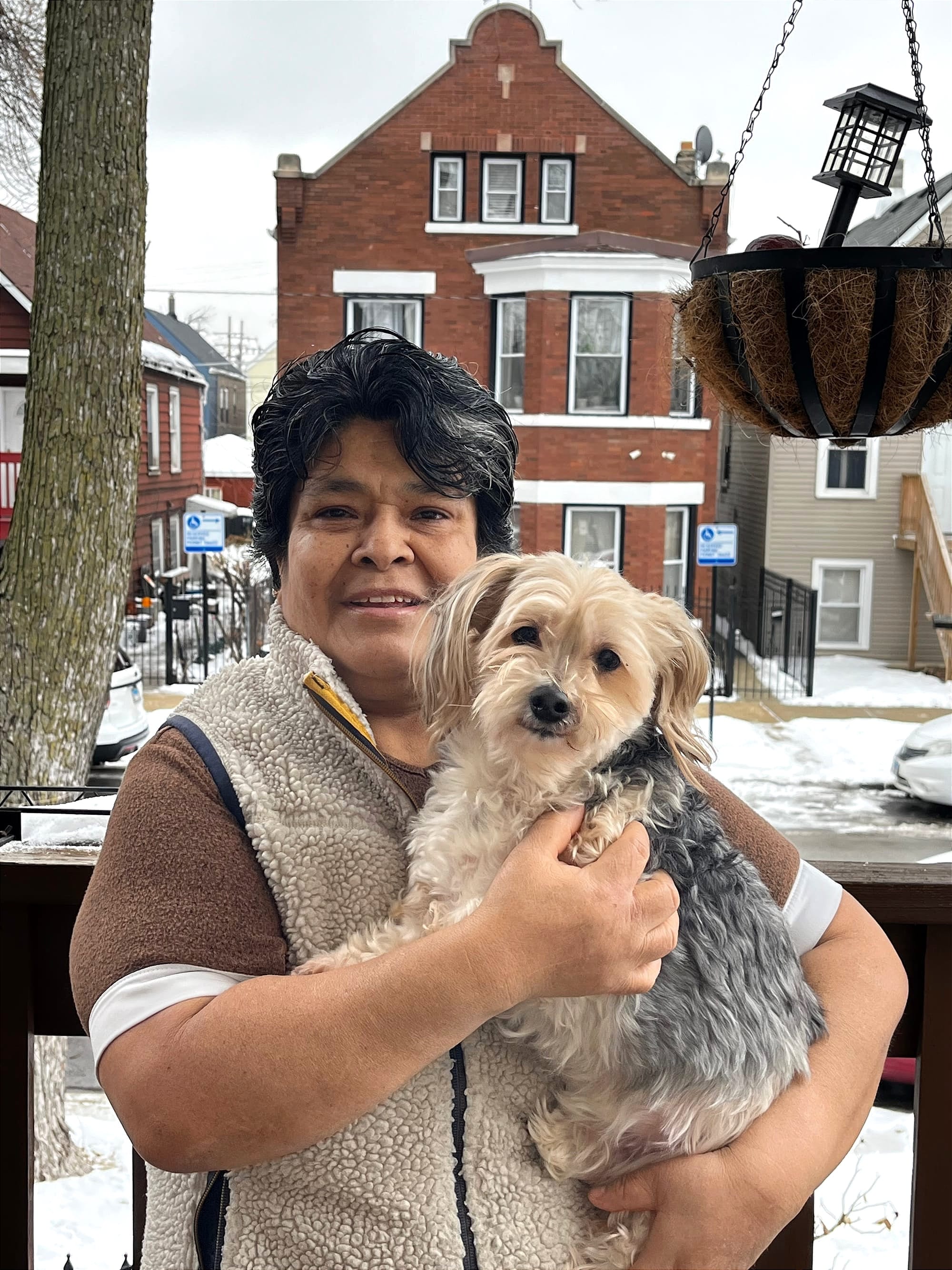 A woman smiles while holding a small gray and white dog on a porch, with a red brick building and snow-covered street visible behind her.